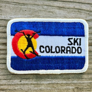 May include: Embroidered patch featuring the Colorado state flag design with a skier silhouette. The patch has a blue background with a white stripe, a red and yellow circle, and the words "SKI COLORADO" in black.