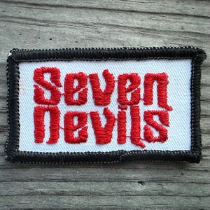 May include: Embroidered patch with the words "Seven Devils" in red thread on a white background. The patch has a black border with a white stitch pattern and a rectangular shape.
