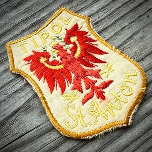 May include: A shield-shaped embroidered patch on a light beige fabric base. It features a red eagle emblem in the center, with the yellow embroidered text TIROL at the top and St. Anton at the bottom. The edges are finished with a yellow border.
