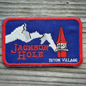 May include: A rectangular embroidered patch with a blue background and a red border. It features white mountain peaks, a red clock tower, and red embroidered text that reads JACKSON HOLE, with white text that reads TETON VILLAGE.