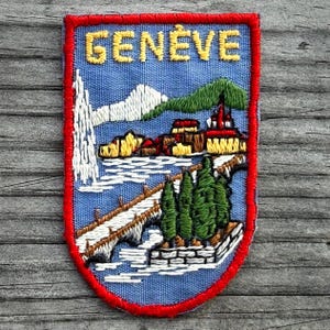 May include: Embroidered patch with a blue background and a red border. The patch features the word "GENÈVE" in yellow, a mountain range, buildings, a bridge, trees, and a waterfall. The patch is a souvenir or travel item.