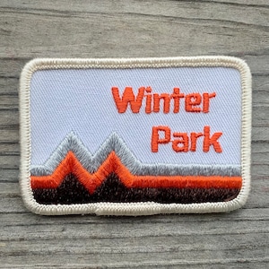 May include: White embroidered patch with orange text that reads "Winter Park". The patch features a stylized mountain range in orange, gray, and brown.