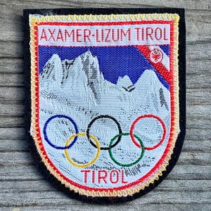 May include: A white and red embroidered patch with the text "Axamer-Lizum Tirol" and "Tirol" in a yellow and black border. The patch features a mountain range and the Olympic rings.