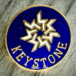 May include: A round, blue and gold Keystone pin. The pin features a white starburst design in the center and the word "KEYSTONE" in gold lettering around the edge. The pin has a gold border and is set against a wooden surface.