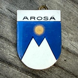 May include: A metal pendant with the word "AROSA" at the top. The pendant features a blue background with a white mountain range and a yellow sun. It has a small gold-colored loop at the top for hanging.