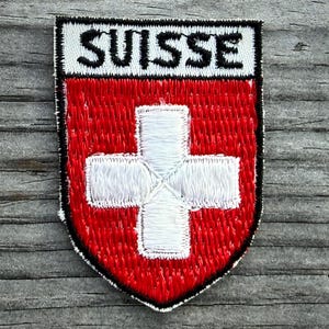 May include: Embroidered patch featuring the word "SUISSE" above a red shield with a white cross. The patch has a black border and is likely a flag emblem or souvenir.