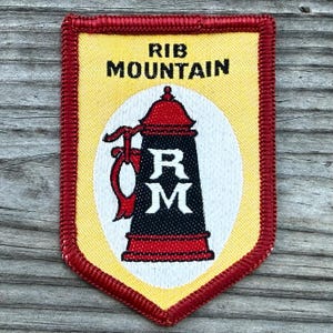 May include: A shield-shaped embroidered patch with a yellow background and a red border. The text RIB MOUNTAIN is stitched in black at the top. A central white oval contains a black and red beer stein graphic with the white letters RM in the center.
