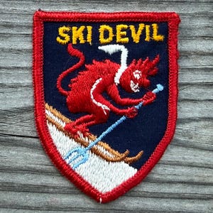 May include: A shield-shaped embroidered patch with a dark blue background and a red border. It features a red devil character skiing on white snow with brown skis, holding a light blue trident pole. The text SKI DEVIL is stitched in yellow.