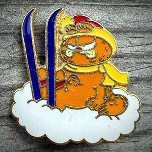 May include: A gold-toned metal enamel pin featuring an orange cartoon cat wearing a yellow hat and scarf, positioned on a white cloud base. The cat is standing next to two tall, dark blue skis with gold outlines, set against a textured background.