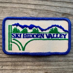 May include: A blue and green embroidered patch with the text "Ski Hidden Valley". The patch features a stylized mountain range and trees.