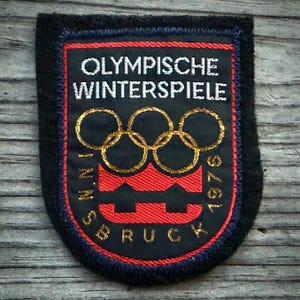 May include: A vintage patch with the text "OLYMPISCHE WINTERSPIELE INNSBRUCK 1976". The patch features the Olympic rings in gold, a red and black design, and a black border. The patch is rectangular with rounded corners.