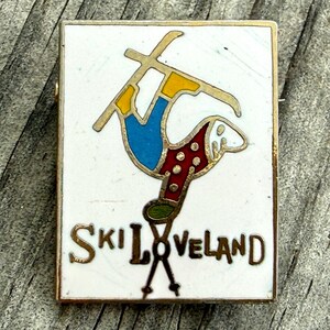 May include: Vintage rectangular pin with a white background and gold trim. The pin features a skier in blue pants, yellow boots, and a red and white polka-dotted jacket. The words "SKI LOVELAND" are written in gold.