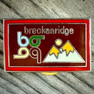 May include: A rectangular pin with a red border and a dark brown center. The pin features the word "breckenridge" in silver lettering, along with a stylized design of mountains, a sun, and the letters "b09".