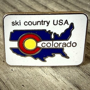 May include: A rectangular pin with a white background features the text "ski country USA" above a blue outline of the United States. The Colorado state flag is in the center, with the word "colorado" below.