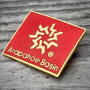 May include: A red square-shaped pin with a gold border and a gold snowflake design. The text "Arapahoe Basin" is written in gold letters below the snowflake.