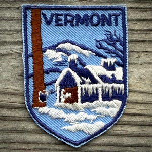 May include: A shield-shaped embroidered patch with a light blue background featuring the word VERMONT in dark blue letters. The design depicts a brown tree trunk, a small cabin covered in white snow and icicles, and dark blue trees.
