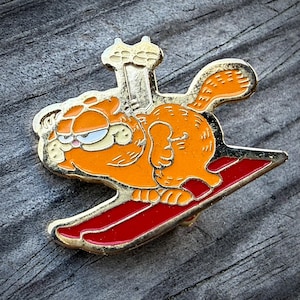 May include: A gold-toned metal enamel pin featuring the orange cartoon cat Garfield skiing on a pair of red skis. The character is depicted in profile with a focused expression, wearing a gold ski hat, with the red skis angled diagonally.