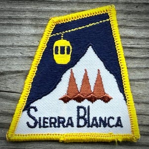May include: A trapezoidal embroidered patch with a bright yellow stitched border. The design features a dark blue sky with a yellow gondola, a white snow-covered mountain peak, three brown pine trees, and the text Sierra Blanca in dark blue.