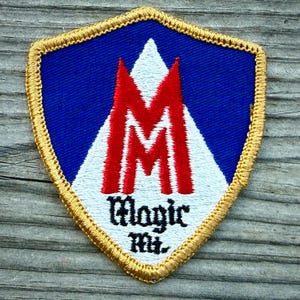 May include: A shield-shaped embroidered patch with a blue background and a gold border. It features a large red letter M centered over a white triangular shape. Below the letter M, the text Magic Mt. is embroidered in black gothic-style lettering.
