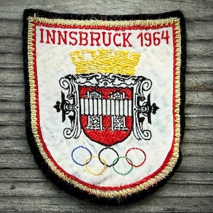 May include: Embroidered patch featuring the text "INNSBRUCK 1964" above a crest and Olympic rings. The patch has a white background with red and gold trim, and a black border. The crest is red and white with a gold crown.