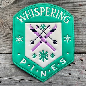 May include: A green and white sign with the words "WHISPERING PINES" and a graphic of crossed skis and snowflakes. The sign is a shield shape and is mounted on a weathered wood surface.