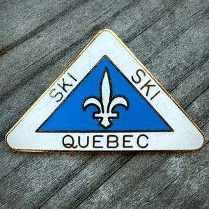 May include: A triangular pin with a gold border, featuring a blue center with a white fleur-de-lis. The words "SKI" and "QUEBEC" are displayed in white text on the pin, which is set against a wooden surface.
