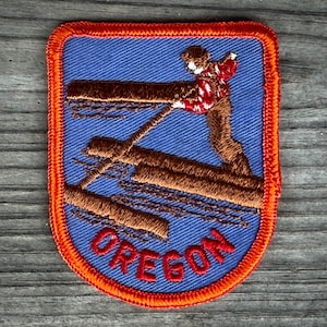 May include: Embroidered patch with a blue background and an orange border. The patch features a lumberjack figure standing on logs, with the word "OREGON" in red letters at the bottom. The lumberjack is wearing a red and white patterned shirt.