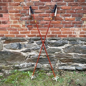 May include: Two wooden ski poles with black tips and white straps, crossed in an X shape against a brick wall background.
