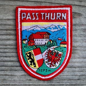 May include: A red, white, and blue embroidered patch with the text "PASS THURN" and a depiction of a mountain village with a red roofed building. The patch also features two shields with crests, one with a red eagle and the other with a black lion. The text "1273 m" is also visible on the patch.