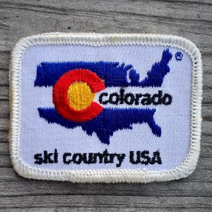 May include: White embroidered patch with a blue and white outline of the United States with the state of Colorado highlighted in red, yellow, and white. The text "Colorado" is embroidered in black and the text "Ski Country USA" is embroidered in black below the outline.
