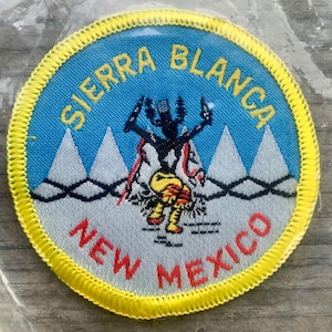 May include: A yellow embroidered patch with a blue background and white mountains. The patch features a black silhouette of a person with a red and yellow headdress, holding two sticks. The text "SIERRA BLANCA NEW MEXICO" is embroidered in yellow and red.