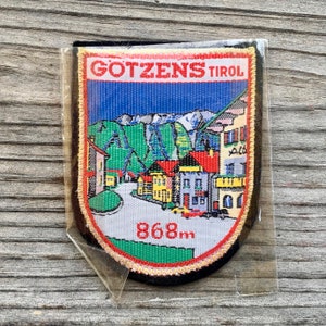 May include: A colorful embroidered patch with a depiction of a village in the Austrian Alps. The patch features a blue sky, green mountains, and a small village with red roofs. The text "GOTZENS TIROL" and "868m" are embroidered on the patch.