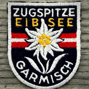 May include: Embroidered patch with the words "ZUGSPITZE EIBSEE GARMISCH" in white and yellow lettering. The patch features a white edelweiss flower with a yellow center, and red and white stripes on a navy blue background.