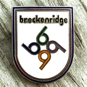May include: A rectangular pin with a white background and the word "breckenridge" in black. The pin features the numbers "6969" in green, purple, orange, and blue. The pin has a silver border with a blue and purple trim.