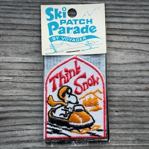 May include: A vintage "Ski Patch Parade" patch with the words "Think Snow" in red script. The patch features a cartoon skier on a snowmobile and a small mountain scene. The patch is on a cardboard backing.