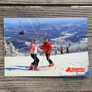 May include: Vintage postcard featuring a ski resort scene. Two skiers in colorful ski suits are on a snowy slope, with a ski lift overhead. The Okemo Mountain Resort logo is in the lower right corner, with a scenic mountain view in the background.