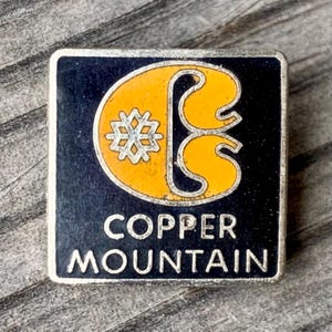 May include: A vintage silver pin with a black background and a yellow stylized letter 'C' with a snowflake inside. The text 'Copper Mountain' is printed in silver below the letter.