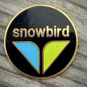 May include: A round black pin with the word "snowbird" in gold lettering. The pin features a blue and green design at the bottom. The pin has a gold border.