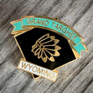 May include: Gold-colored pin with a black diamond shape featuring a Native American headdress design. The pin has a turquoise blue ribbon with the text "GRAND TARGHEE WYOMING" in gold lettering.