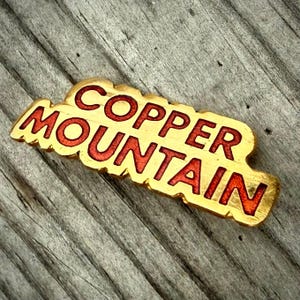 May include: A gold-colored, rectangular pin with the words "COPPER MOUNTAIN" in red, stylized lettering. The pin has a textured, irregular border. The background is a weathered, gray wooden surface.