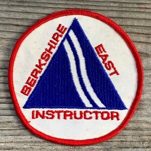May include: White and red circular patch with a blue triangle in the center. The triangle has a white stripe down the center. The text "Berkshire East Instructor" is embroidered in red around the triangle.