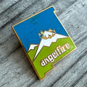 May include: A rectangular enamel pin with a gold border. The pin features a blue sky, white mountains, and green foreground with the word "angelfire" in white. The pin is set against a weathered wooden surface.
