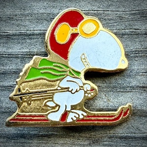 May include: A gold-toned metal enamel pin featuring a white cartoon dog wearing a red aviator cap with yellow goggles and a green scarf. The character is depicted in a skiing pose, holding a ski pole while wearing red skis on a gold-colored base.