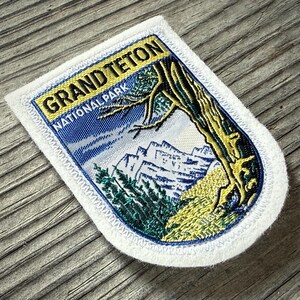May include: A rectangular patch with a white border featuring the text "GRAND TETON NATIONAL PARK." The patch depicts a mountain range, trees, and a blue sky. The patch is made of fabric and has a textured appearance.