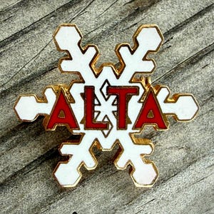 May include: A snowflake-shaped pin with a gold border, white enamel, and the word "ALTA" in red letters. The pin is set against a wooden background. This winter-themed pin is a decorative accessory.