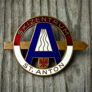 May include: A circular metal pin featuring a blue letter A with a white wavy line design inside. The border is red and white with the text SKIZENTRUM ST. ANTON. A horizontal gold-toned bar extends behind the circle, and a small red eagle emblem sits at the base of the letter A.