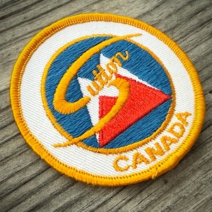 May include: Embroidered patch with a circular design. The patch features a blue background with a red and white mountain graphic. The text "Wilson" is in gold, along with the word "CANADA" around the border. The patch has a gold border.