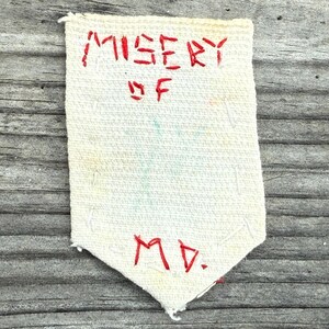 May include: Cream-colored, rectangular fabric patch with red embroidered text. The text reads "MISERY OF MD." The patch has a pointed bottom and is placed on a weathered, gray wooden surface.