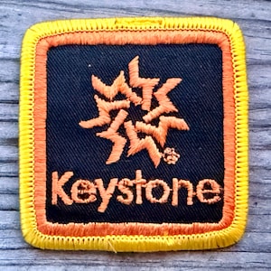 May include: Square patch with a black background, orange border, and yellow stitching. The patch features an orange starburst design and the word "Keystone" in orange text.