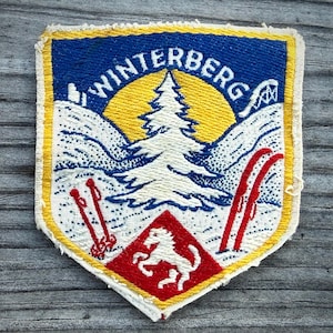 May include: Vintage Winterberg patch with a shield shape. The patch features a blue sky with the word "WINTERBERG" in white, a yellow sun, a white pine tree, and red skis. A red diamond with a white horse is at the bottom.